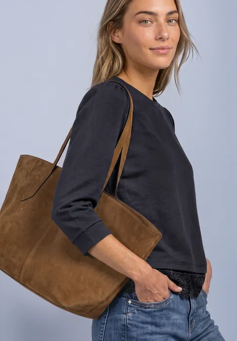 Frau in schwarzem Dreiviertelshirt und Jeans, trägt eine braune Wildledertasche über der Schulter.