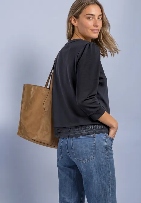 Frau in schwarzer Bluse, Jeans und brauner Ledertasche über der Schulter, blickt seitlich in die Kamera.