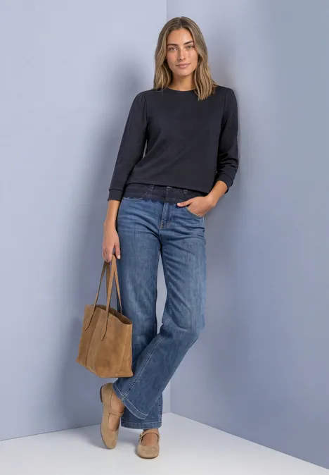 Frau lehnt an grauer Wand, trägt schwarzen Pullover, blaue Jeans, beigen Loafer und braune Tasche.