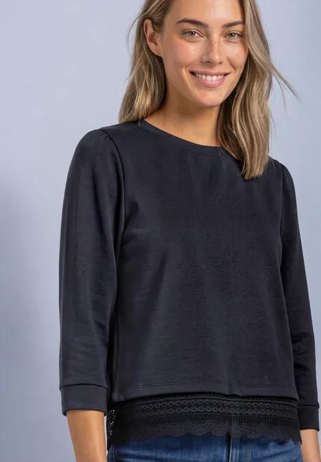 Frau trägt lächelnd schwarzes Shirt mit 3/4-Ärmeln und Spitzenbesatz vor hellem Hintergrund.