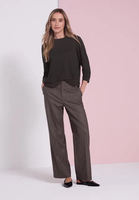 Frau in schwarzer Bluse und weiten grauen Hosen, schwarze Ballerinas, vor hellem rosa Hintergrund.