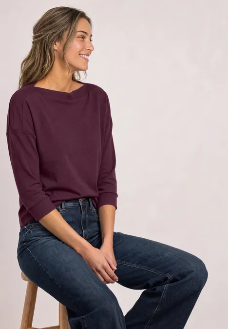 3/4-Arm Shirt mit Rippstrukturdetails rustic rouge Lächelnde Frau im weinroten Shirt und Jeans sitzt seitlich auf einem Hocker vor hellem Hintergrund.