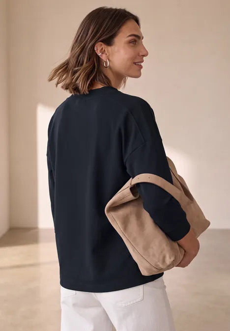 Frau in schwarzem Pullover und weißer Hose hält eine beige Tasche vor minimalistisch hellem Hintergrund.
