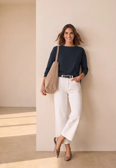 Eine Frau lehnt an einer beigen Wand, trägt schwarzen Pullover, weiße Culottes, beige Tasche und Schuhe, lächelt.