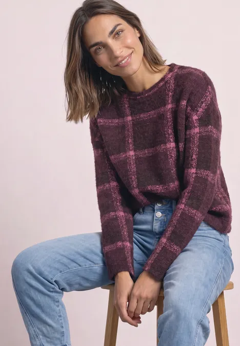 Vrouw zit op kruk, draagt bordeauxrood geruit trui en blauwe jeans, licht glimlachend.
