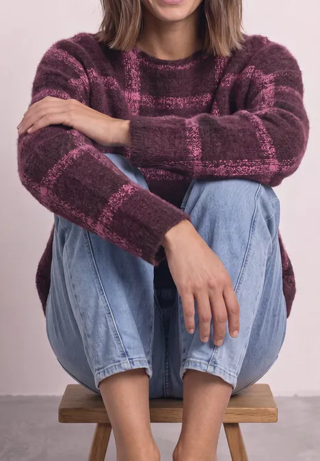 Vrouw zit op een houten kruk, draagt een paars-roze geruit vest en blauwe jeans, met armen om haar knieën.