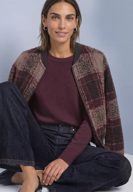 Vrouw in geruit jack en paarse trui zit ontspannen in donkere jeans voor een egale achtergrond.