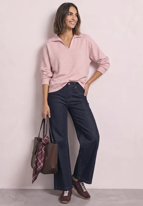 Frau trägt rosa Pullover und weite Jeans, hält eine braune Ledertasche mit kariertem Schal.