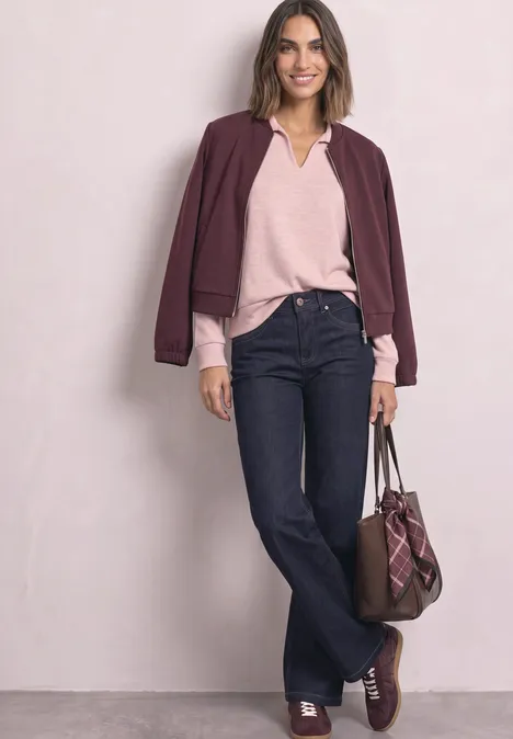 Frau in lässig-elegantem Outfit: bordeauxfarbene Jacke, rosafarbener Pullover, dunkelblaue Jeans, Tasche und Schuhe in Braun.