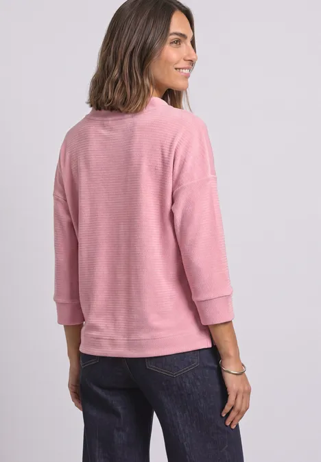 Frau, die ein rosa Sweatshirt mit 3/4-Ärmeln und dunkle Jeans trägt, schaut über die Schulter.