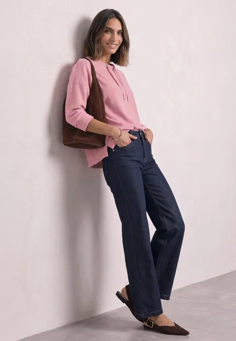 Frau vor heller Wand, trägt rosa Pullover, weite blaue Hose und braune Tasche, lächelt.