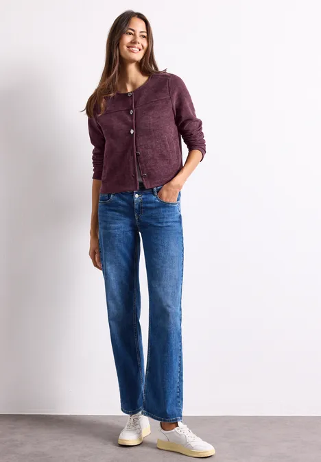 Une femme souriante porte un gilet violet, un jean large et des baskets blanches sur fond clair.