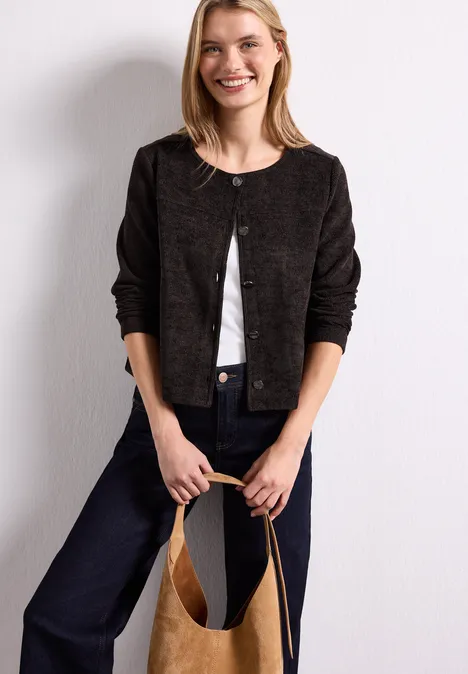 Jas met lange mouwen, ronde hals en knopen black coffee Frau lehnt an weißer Wand, trägt schwarzen Cardigan, weiße Bluse, Jeans und hält eine beige Tasche.