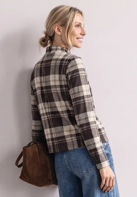 Frau in kariertem Blazer, Jeans und brauner Tasche, lächelnd seitlich nach rechts gewandt.