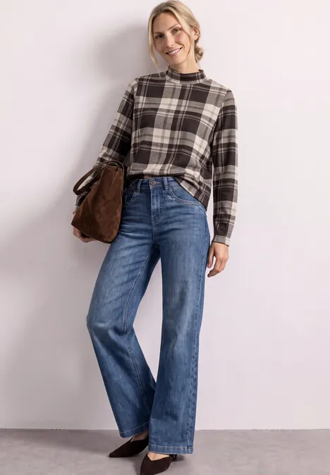 Eine lächelnde Frau trägt eine karierte Bluse, weite Jeans und eine Umhängetasche vor hellem Hintergrund.