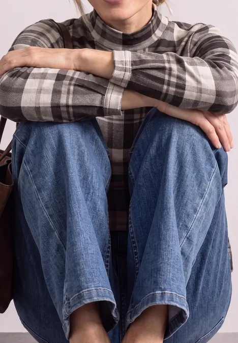 Eine Person sitzt mit verschränkten Armen auf den Knien, trägt eine karierte Bluse und Jeans.