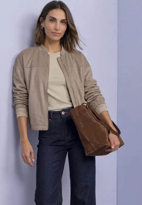 Frau in beiger Bomberjacke, hellem Shirt, dunklen Jeans hält braune Wildledertasche vor lila Wand.