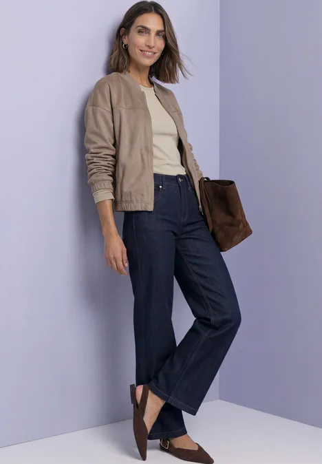 Frau in taupefarbener Bomberjacke, beigem Top, dunkler Jeans und braunen Loafers vor lila Wand.