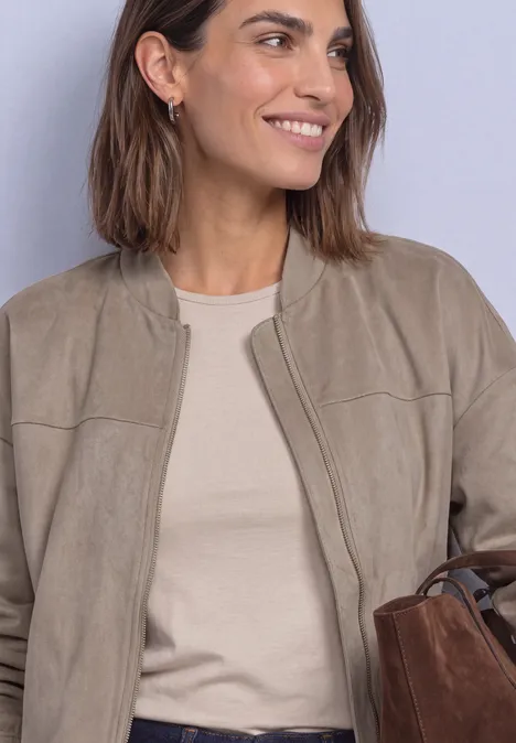 Eine lächelnde Frau trägt eine hellbraune Jacke, beigefarbenes Shirt und hält eine braune Tasche.