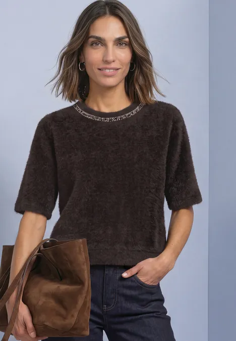 Frau mit braunem, samtigem Kurzarmshirt, dunklen Jeans und brauner Wildledertasche vor neutralem Hintergrund.