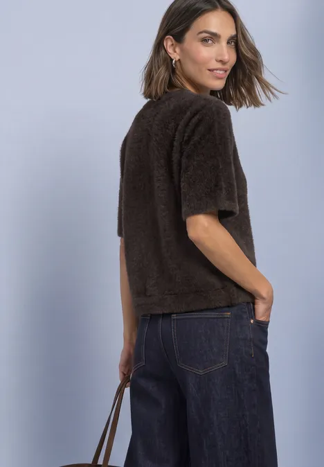 Frau im Profil trägt kurzärmligen braunen, flauschigen Pullover, dunkelblaue Jeans und hält eine Tasche.