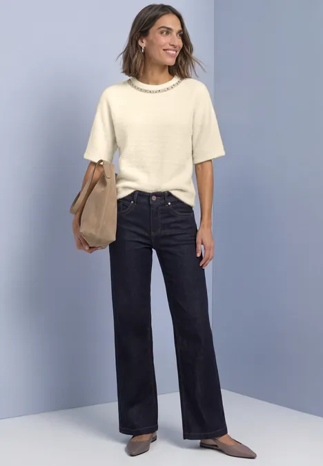 T-shirt à col rond orné de strass pure beige Femme en top court crème en maille, jean évasé foncé, chaussures marron et sac, devant un fond bleu