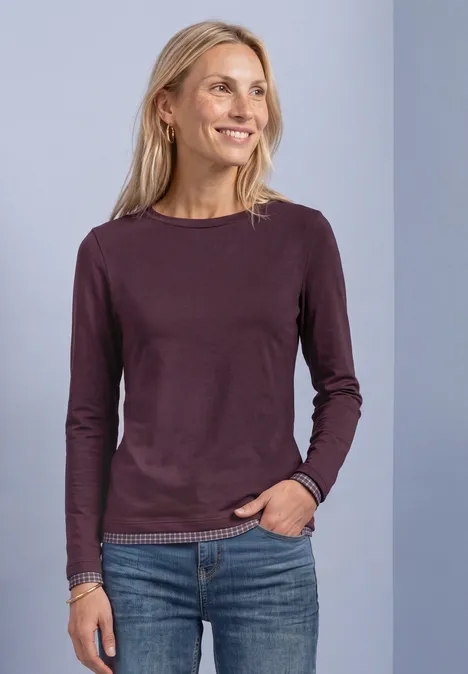 Lächelnde blonde Frau in lila Langarmshirt und Jeans, eine Hand in der Tasche, seitlich schauend.