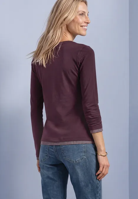Eine lächelnde Frau von hinten, trägt lila Langarmshirt und blaue Jeans vor hellem Hintergrund.