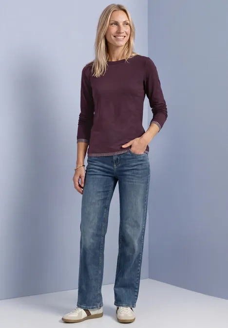 Eine Frau in Jeans, beerenfarbenem Pullover und weißen Sneakern vor hellem, bläulichem Hintergrund.