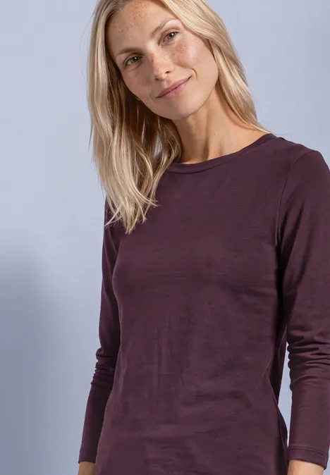 Eine lächelnde blonde Frau in plumafarbenem Langarmshirt vor helllila Hintergrund.