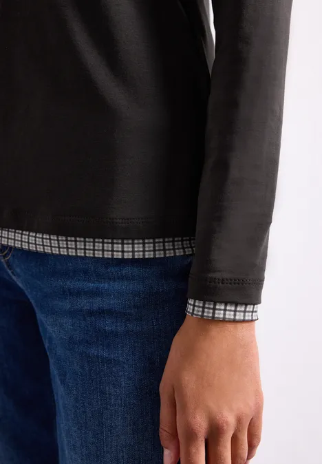 Teilansicht einer Person mit schwarzem Langarmshirt über kariertem Hemd und blauer Jeans.