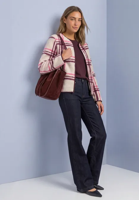 Vrouw in wijd uitlopende jeans, geruite jas en grote rode tas voor een lichtblauwe achtergrond.