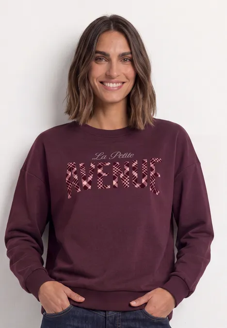 Frau in bordeauxrotem Pullover mit Blütenstickerei und Schriftzug.