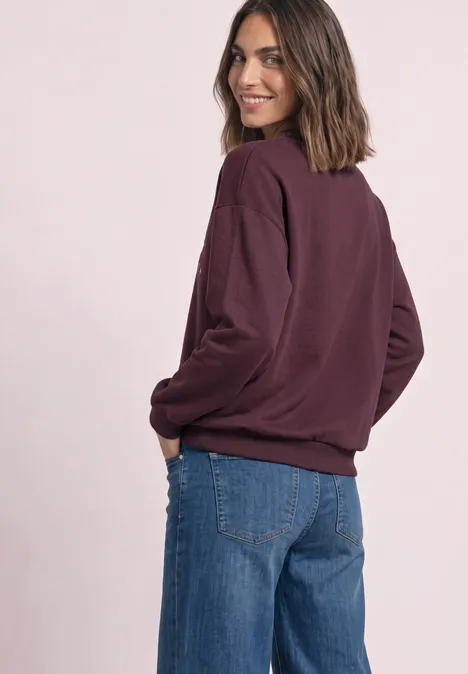 Frau mit weinrotem Pullover und blauen Jeans, lächelnd über die Schulter blickend.