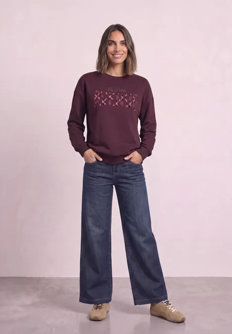 Frau im bordeauxfarbenen Sweatshirt mit Herzmuster, weiter Jeans und beigen Sneakern vor hellem Hintergrund.