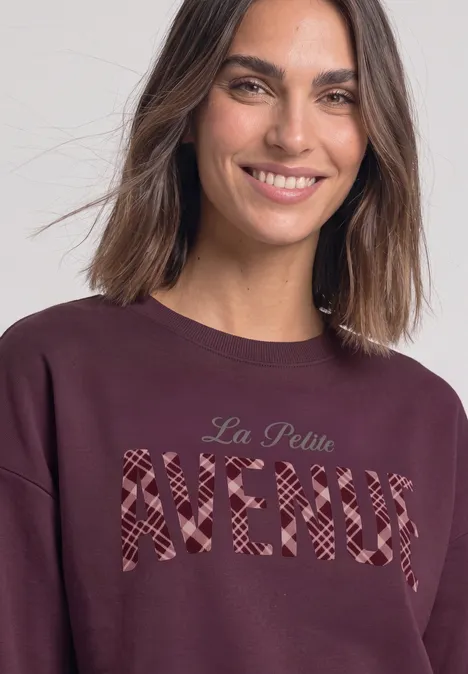 Lächelnde junge Frau in bordeauxrotem Sweatshirt mit Schriftzug „La Petite AVENUE“.