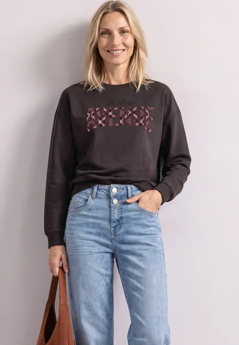 Eine lächelnde Frau trägt schwarzen Pullover mit rosa Kreuzmotiv, hellblaue Jeans und hält eine braune Tasche.