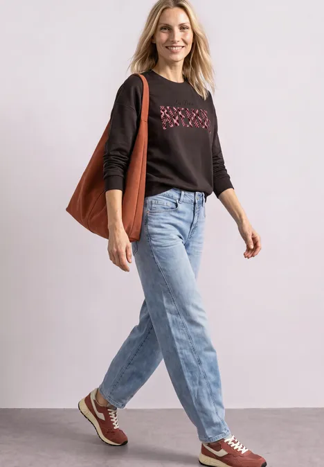 Frau in Jeans, schwarzem Pullover und Sneakern, trägt eine braune Tasche und läuft lächelnd.