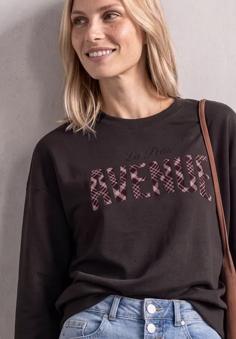 Eine lächelnde Frau trägt einen schwarzen Pullover mit zebrastreifenem Schriftzug "AVENUE" und blaue Jeans.