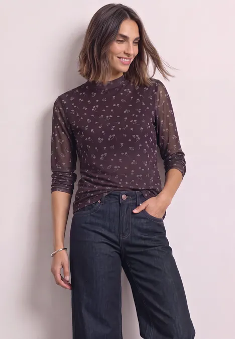 Frau mit halblangem dunklem, gemustertem Oberteil und dunklen Jeans, die lächelnd in die Ferne schaut.