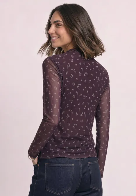 Frau in Jeans und transparentem dunkelrotem Langarmshirt mit kleinem Blumenmuster, lächelnd seitlich stehend.