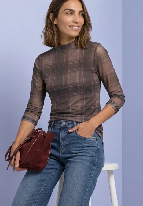 Frau in kariertem Langarmshirt und Jeans, hält eine dunkelrote Tasche vor hellem Hintergrund.