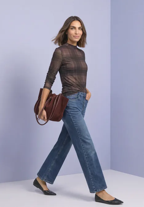 Frau in karierter Bluse, weiter Jeans und Ballerinas mit brauner Ledertasche vor pastellfarbenem Hintergrund.