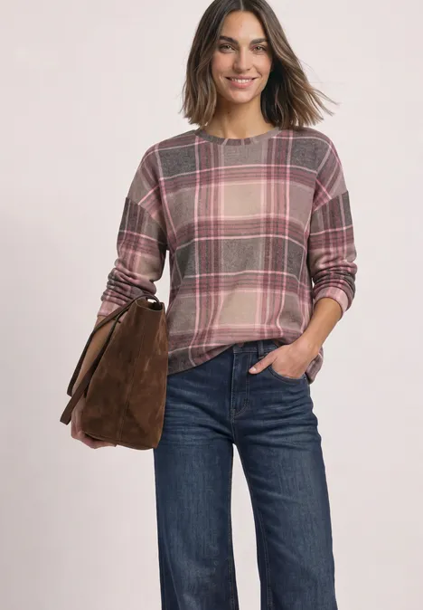 Eine lächelnde Frau trägt einen karierten Pullover, blaue Jeans und eine braune Tasche.