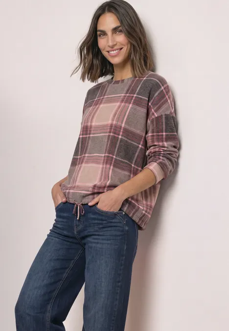 Eine lächelnde Frau in einem karierten Longsleeve und Jeans, die Hände in den Taschen.
