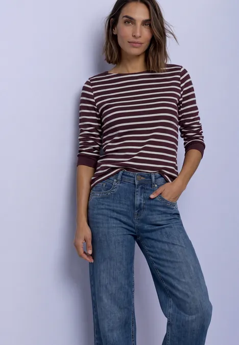 Eine Frau trägt ein gestreiftes Langarmshirt und blaue Jeans, steht lässig mit einer Hand in der Hosentasche.