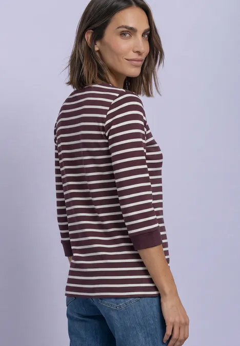 Eine Frau in lila-weiß gestreiftem Langarmshirt und Jeans vor hellem violettem Hintergrund.