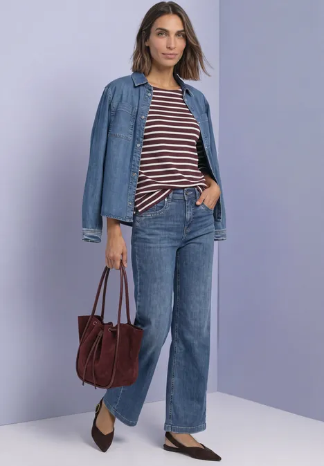 Frau vor lila Wand in weiten Jeans, gestreiftem Shirt, offener Jeansjacke, braunen Pumps und roter Tasche.