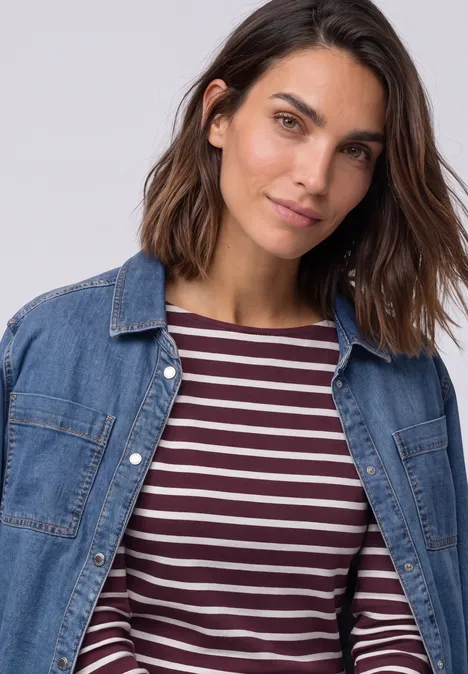 Frau mit schulterlangem braunem Haar trägt blaues Jeanshemd über gestreiftem Shirt.