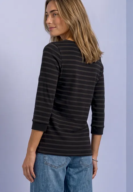 Eine Frau in Jeans und schwarz-grau gestreiftem 3/4-Shirt schaut über die Schulter vor hellem Hintergrund.